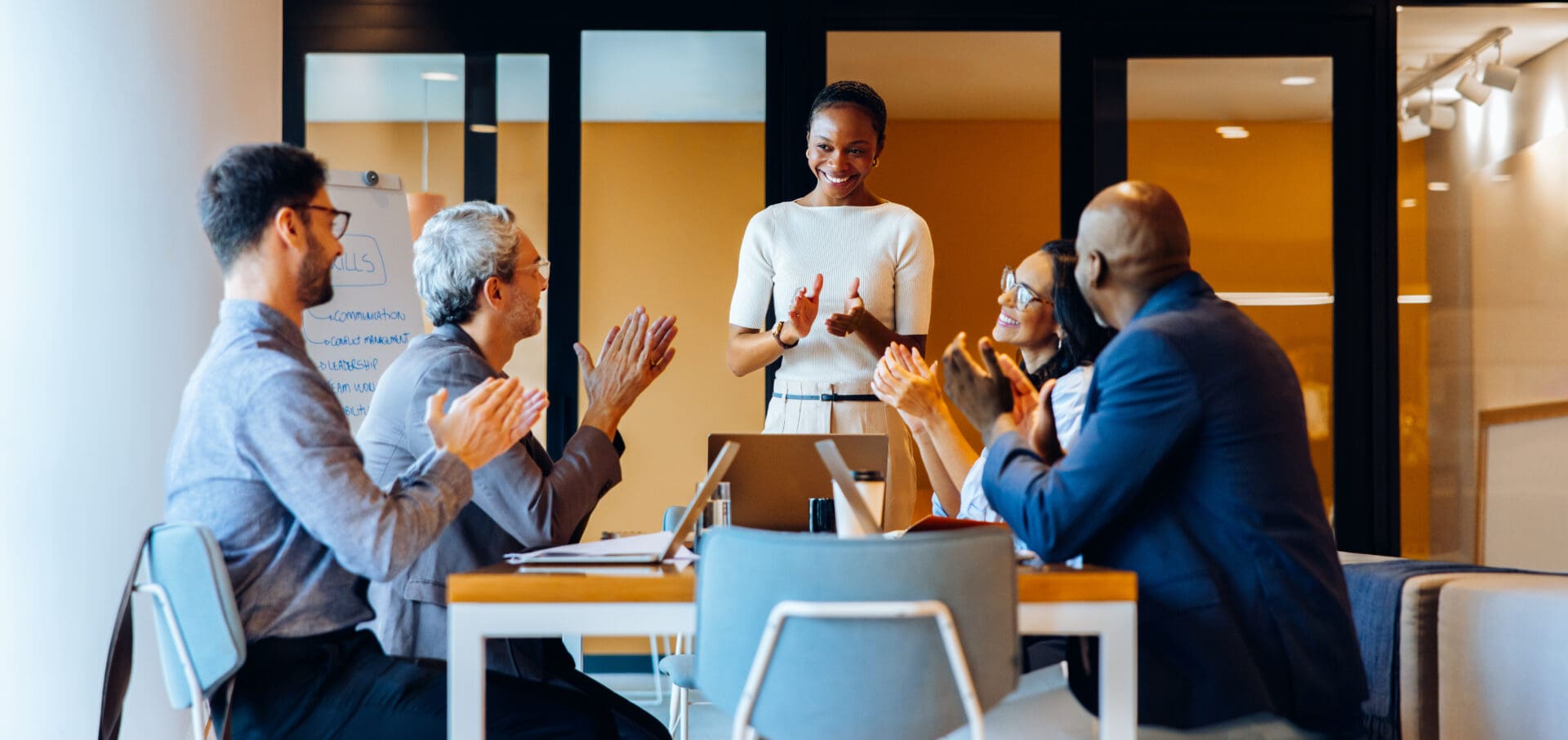 A group of five professionals, men and women, engaging in a lively discussion and applauding an individual standing and presenting at the head of the table in a colorful, welcoming office.
