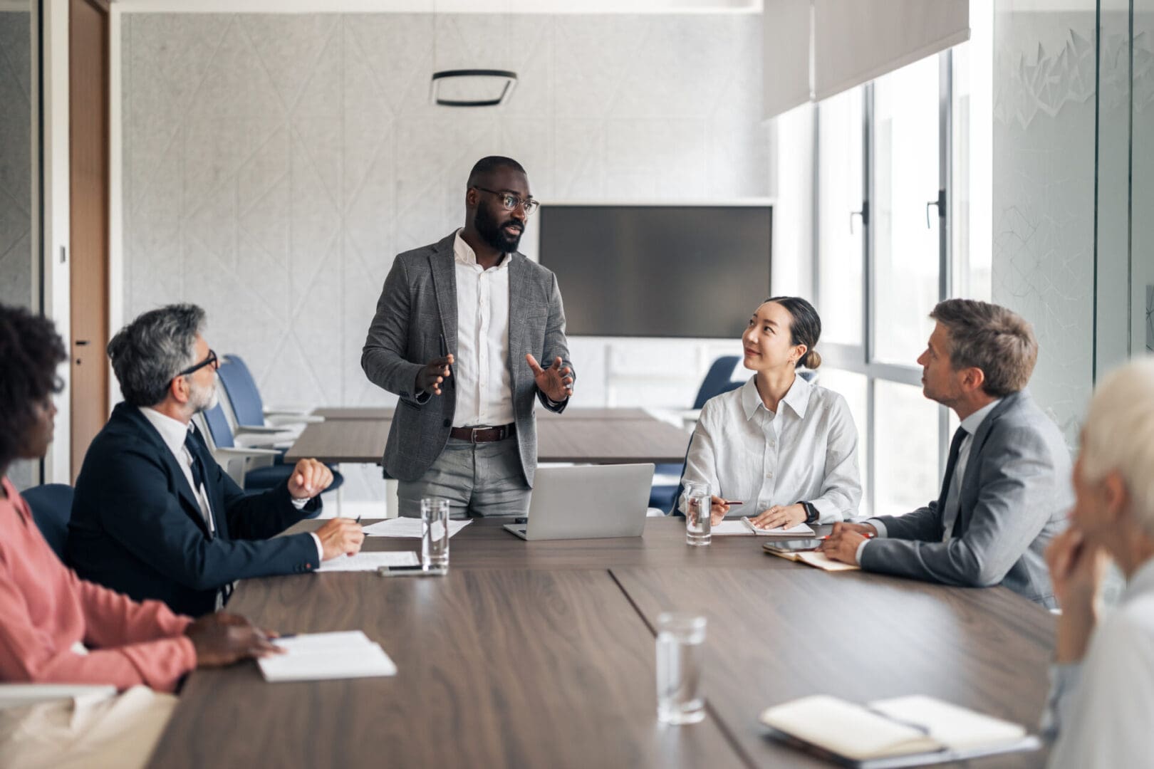 Black businessman presenting strategy to a diverse team in a modern boardroom, colleagues listening attentively around a conference table with laptop and documents