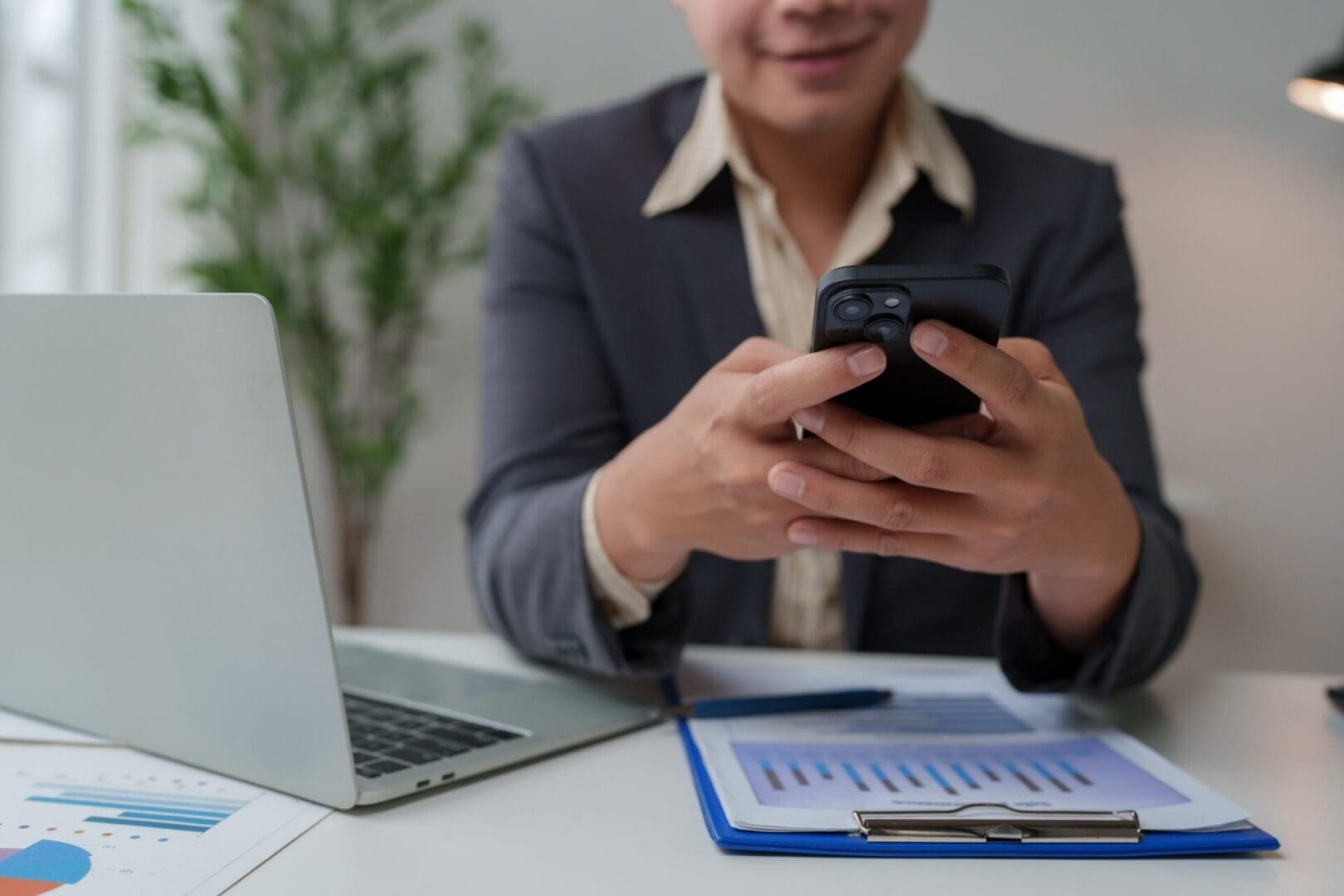 Business person using smartphone and laptop, analyzing financial documents in an office setting