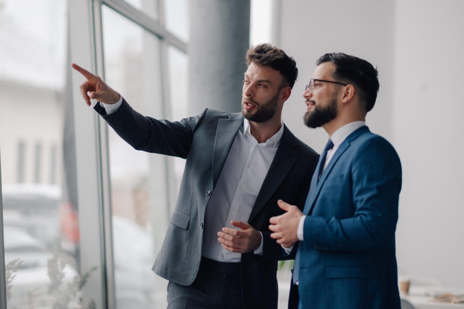 Two young businessmen are discussing strategy while looking out the office window, with one pointing towards something outside