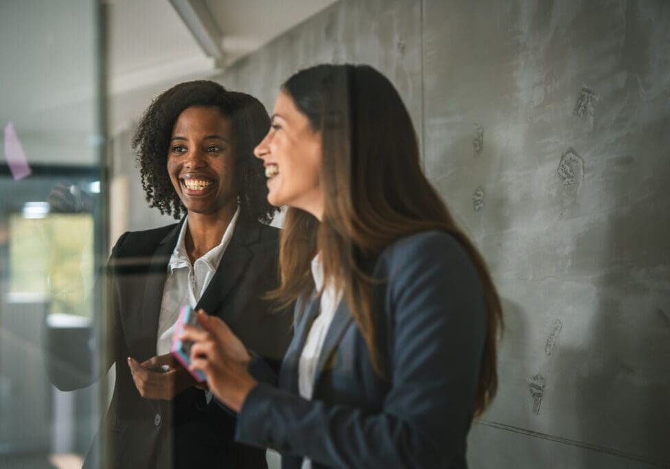 Two women discussing ideas on glass board.