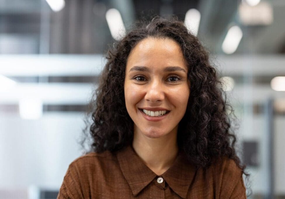 Young curly-haired businesswoman smiling confidently at camera in a modern office headshot, conveying positivity, professionalism and successful leadership in a casual corporate setting