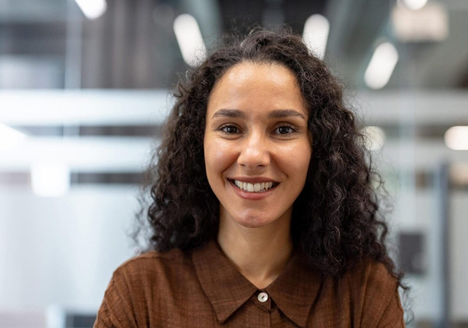Young curly-haired businesswoman smiling confidently at camera in a modern office headshot, conveying positivity, professionalism and successful leadership in a casual corporate setting