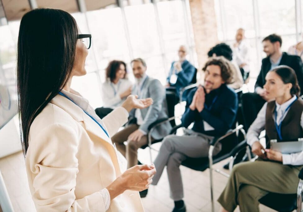 Professional office setting features a female presenter engaging with colleagues. Highlighting teamwork, collaboration, and professional development, this conference environment fosters workplace learning and communication among diverse professionals.
