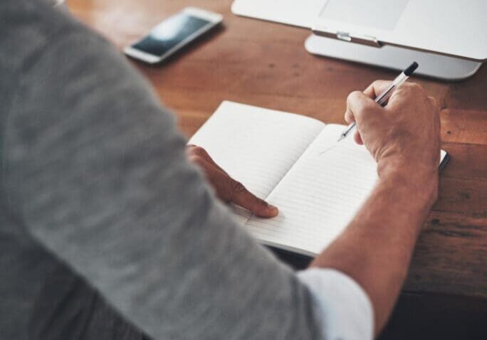 Person writing in notebook beside laptop