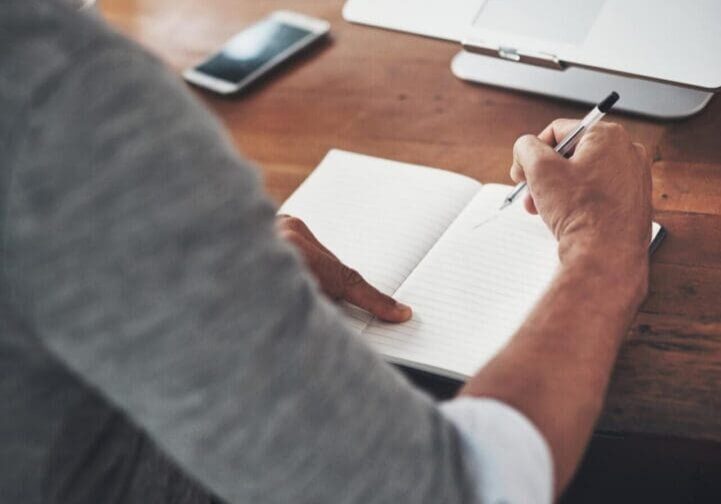 Person writing in notebook beside laptop