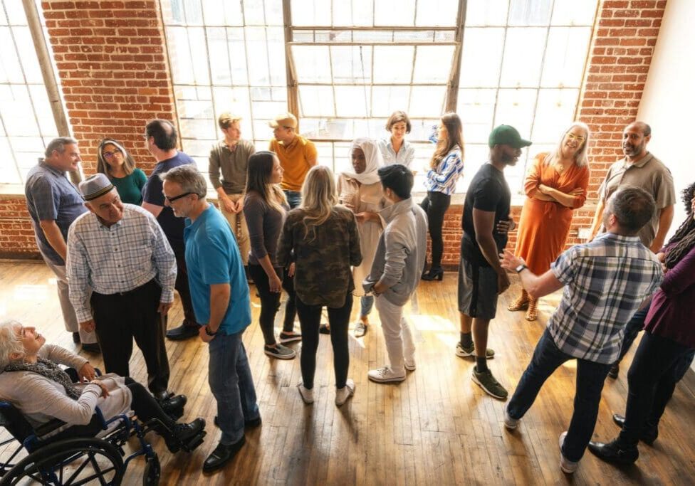 Diverse group of people in a bright room, engaging in conversation. Men and women of various ethnicities, standing and interacting, creating a lively atmosphere. Diverse crowd mingle at social event.