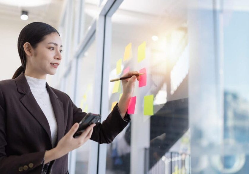 Brainstorming and at idea notes concept, Business woman looking at sticky note on glass wall and sharing ideas in the office.
