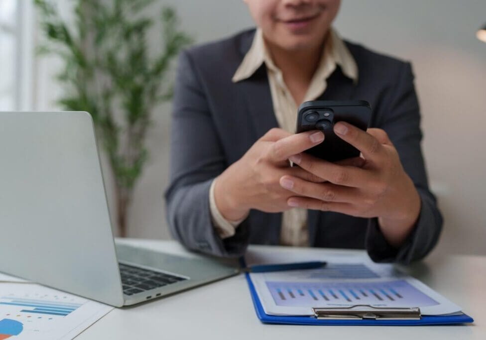 Business person using smartphone and laptop, analyzing financial documents in an office setting