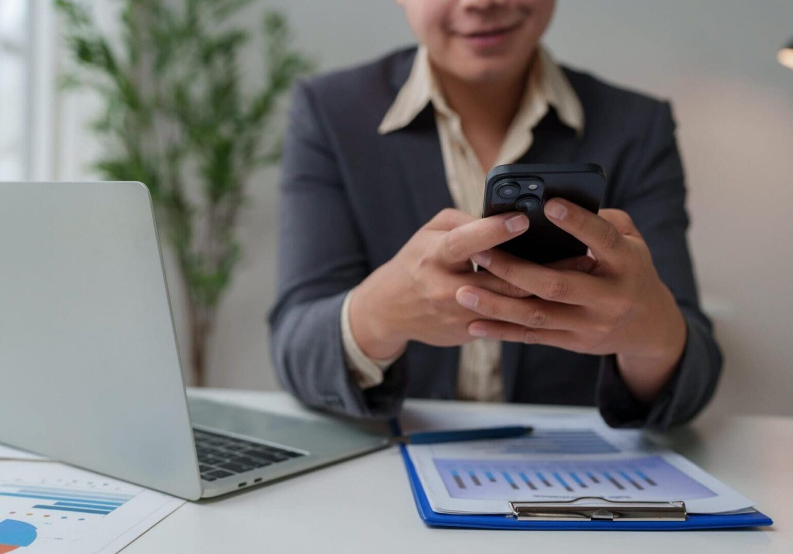 Business person using smartphone and laptop, analyzing financial documents in an office setting