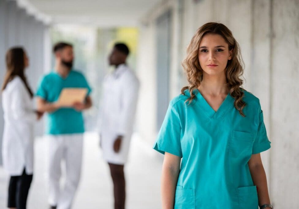Female doctor or nurse in teal scrubs posing at a modern hospital, with colleagues talking in the background
