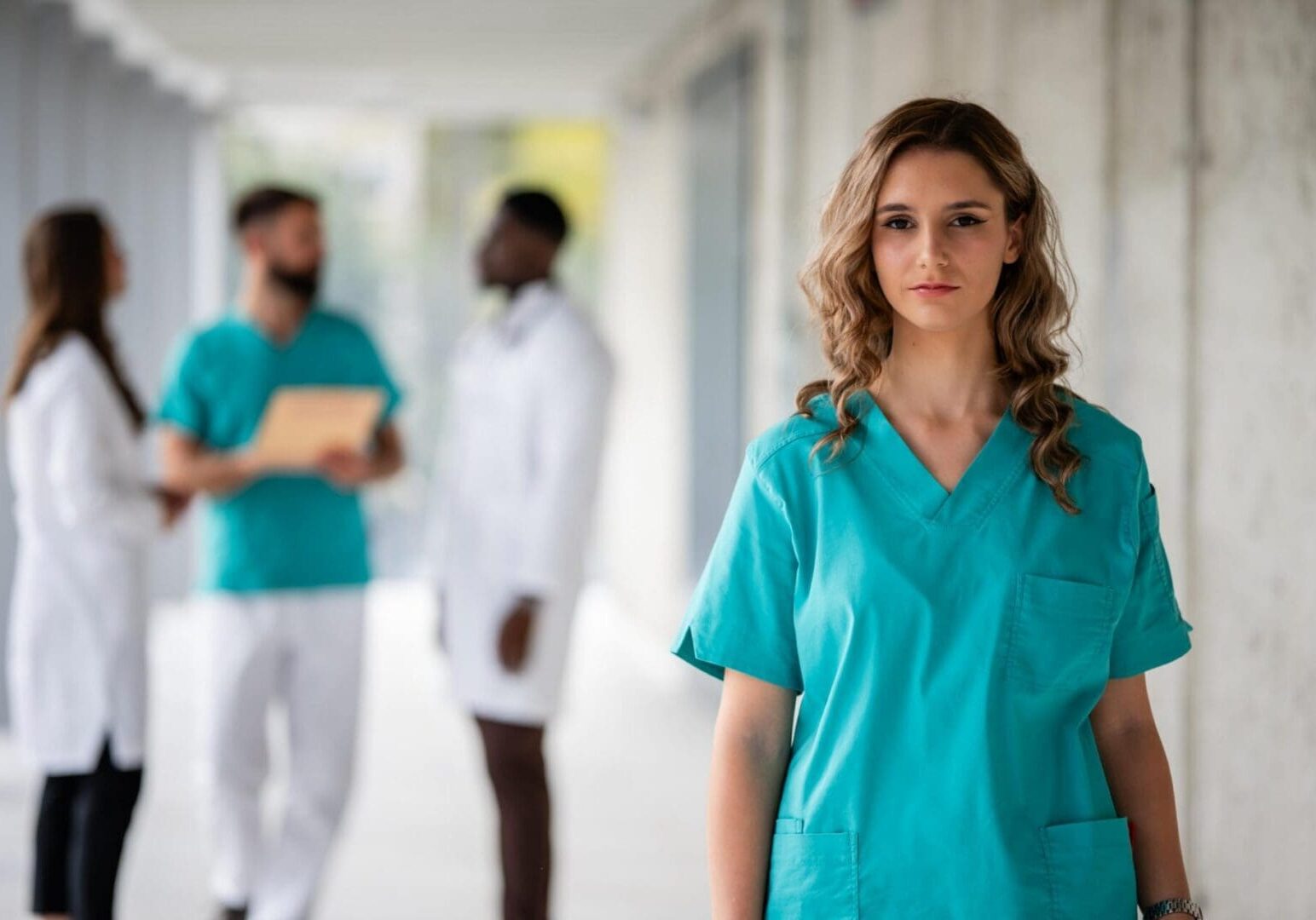 Female doctor or nurse in teal scrubs posing at a modern hospital, with colleagues talking in the background