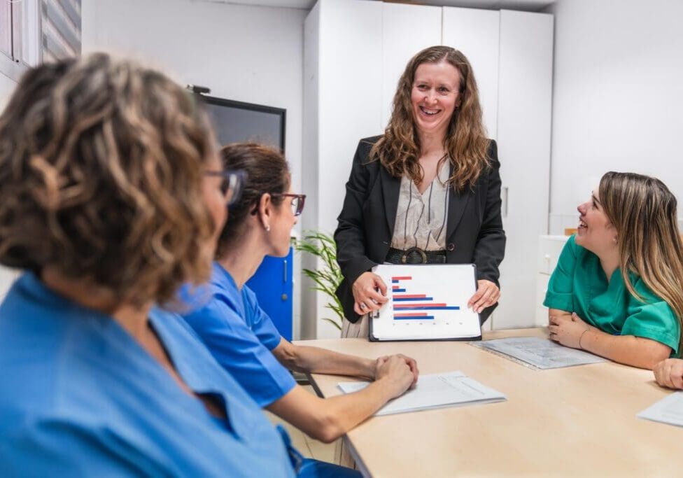 Healthcare professionals in a nursing home setting discuss patient care strategies. A leader presents data on a tablet, fostering collaboration and teamwork