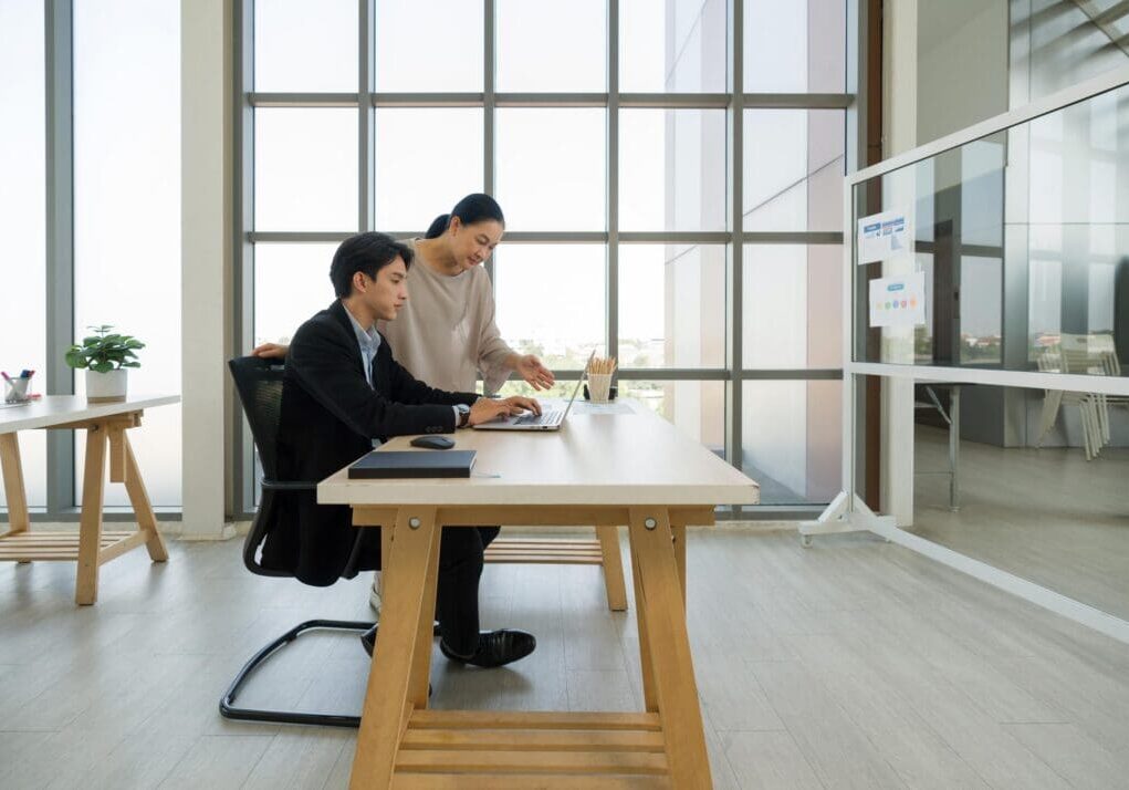 Young man in black suit working on a laptop computer. A senior female colleague or supervisor is pointing at the laptop screen, providing guidance or discussing work-related matters.