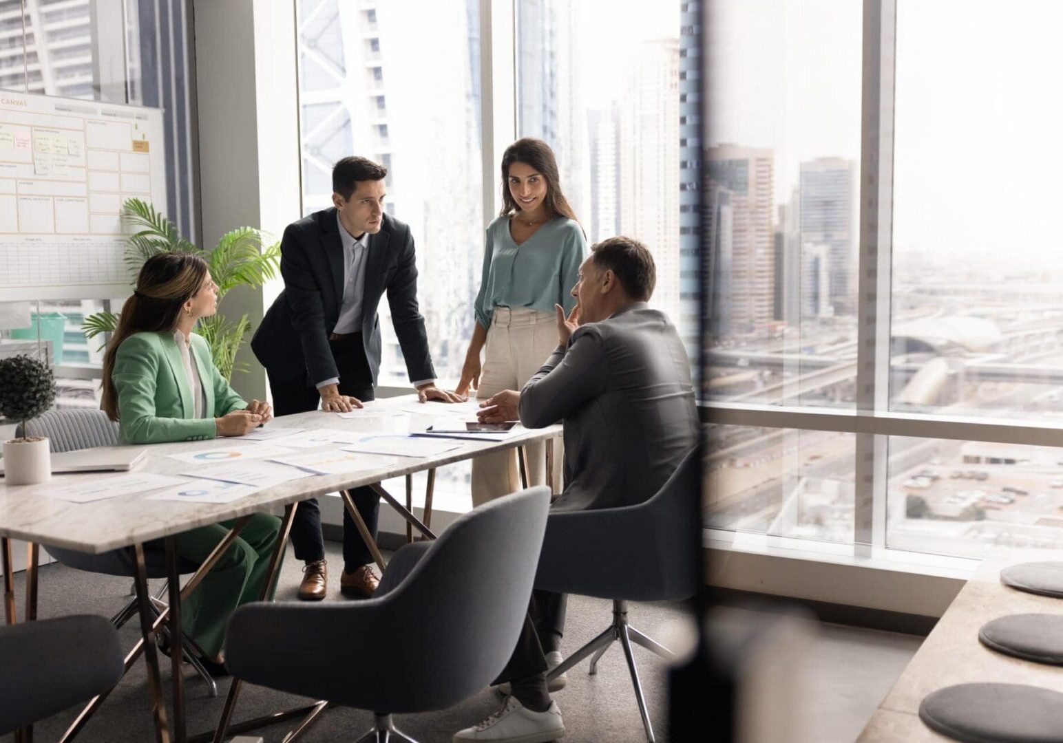 Group of four professionals engaged in srategic business planning meeting in high-rise office with panoramic city view. Team brainstorming ideas, making important decisions. Seminar, briefing event
