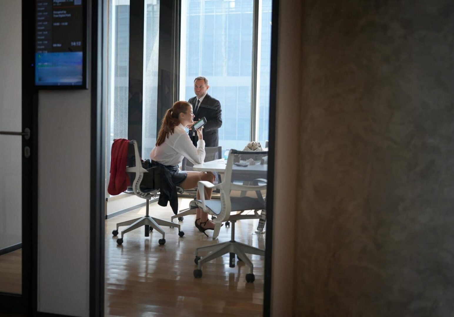 A man in a suit and a woman in business attire engage in a conversation while looking at a mobile device in a bright, contemporary office space