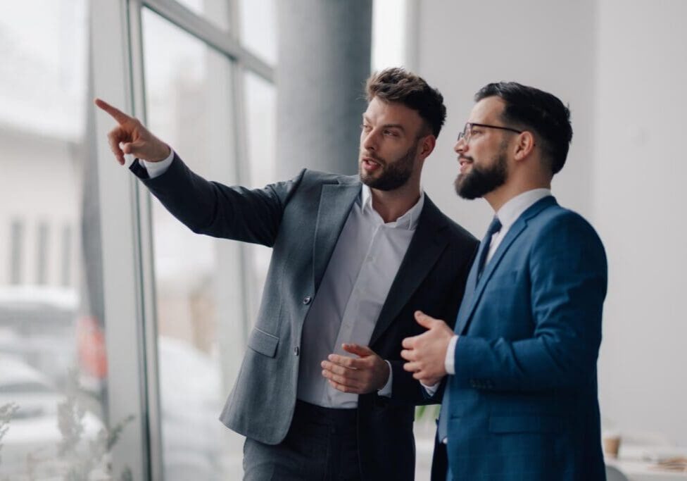 Two young businessmen are discussing strategy while looking out the office window, with one pointing towards something outside