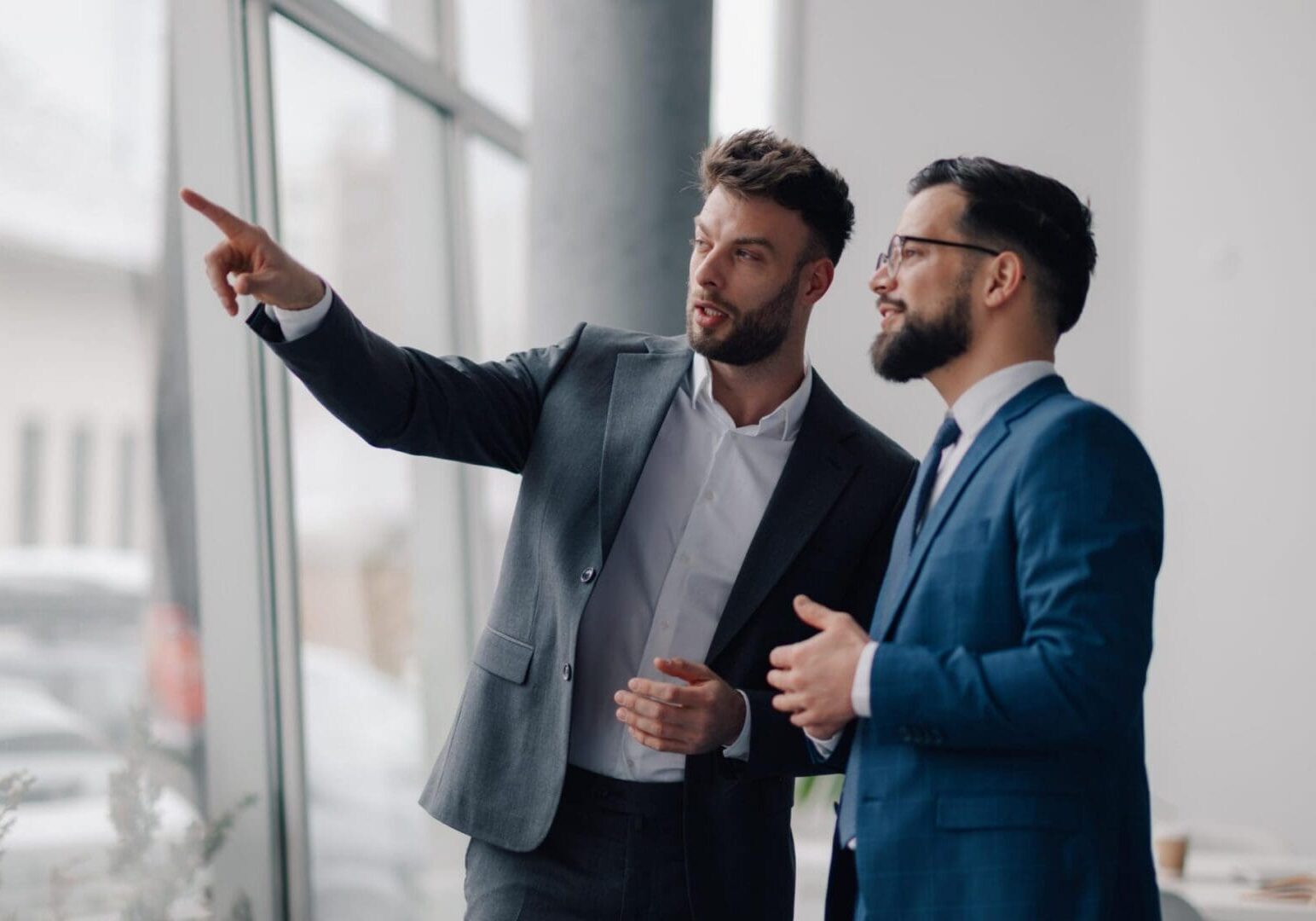 Two young businessmen are discussing strategy while looking out the office window, with one pointing towards something outside